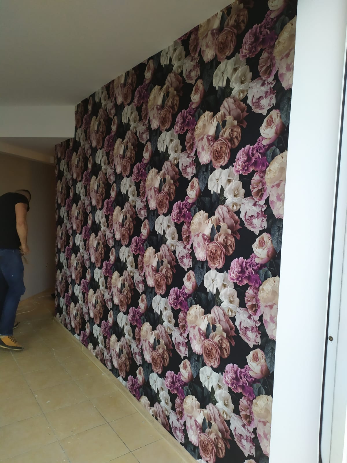 Montaż tapety w kwiaty na ścianie korytarza, widoczny fragment sylwetki montera w roboczych butach.