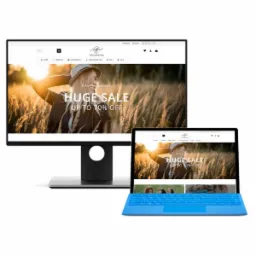 Ekran monitora i laptopa wyświetlają stronę internetową sklepu odzieżowego z promocją do 70% z kobietą w kapeluszu na tle pola pszenicy w promieniach zachodzącego słońca.