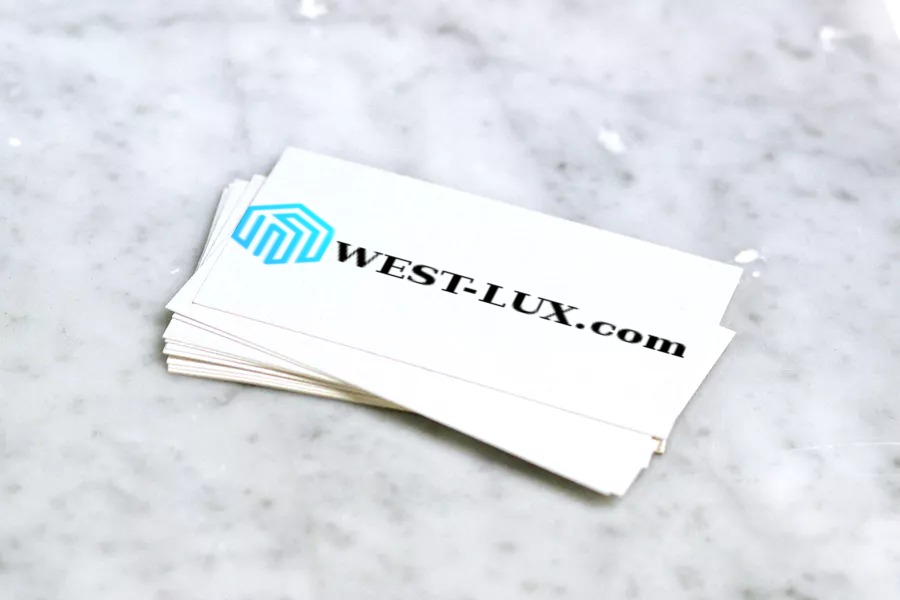 Stos wizytówek z niebieskim logo w kształcie sześcianu i czarnym napisem WEST-LUX.com, leżący na marmurowym tle.