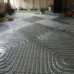 Białe rury ogrzewania podłogowego ułożone w spiralny wzór na metalowej siatce, widoczne elementy konstrukcyjne budynku w tle.