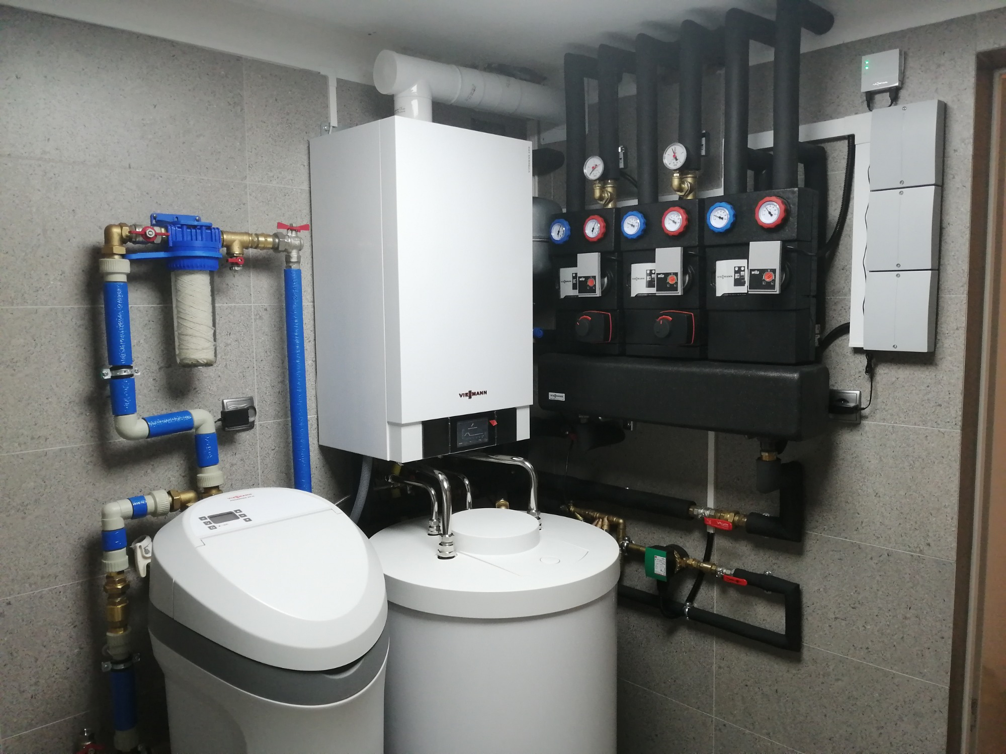 Biały piec grzewczy Viessmann z podłączonymi rurami i manometrami, filtr wody z niebieskimi rurami, zmiękczacz wody, szare płytki na ścianie.
