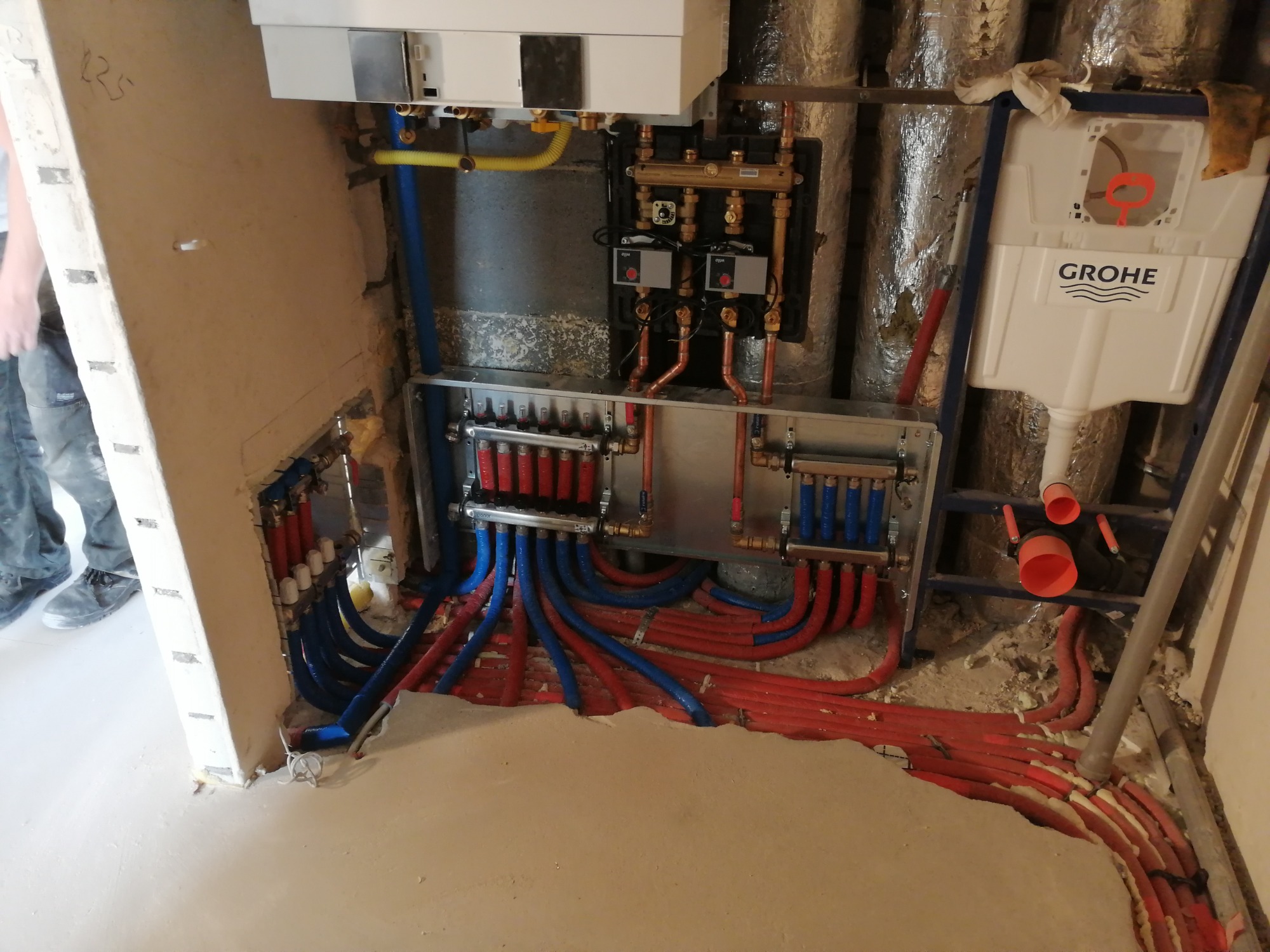 Rozdzielacz ogrzewania podłogowego z rurami czerwonymi i niebieskimi w trakcie montażu, widoczny również piec gazowy i stelaż podtynkowy WC Grohe.