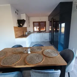 Nowoczesna kuchnia z białymi szafkami i drewnianym blatem, czarne, lakierowane szafy w zabudowie, stół z plecionymi podkładkami i aksamitnymi krzesłami w odcieniach szarości i błękitu.