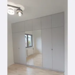 Szara szafa wnękowa z lustrami na całej wysokości, w nowym mieszkaniu z drewnianą podłogą i widokiem na zewnątrz przez okno odbijające się w lustrach.