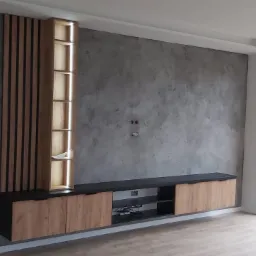 Nowoczesna ściana telewizyjna z szarą fakturą betonu, drewnianymi lamelami, podświetlaną półką i szafką RTV w minimalistycznym salonie z dużym oknem.