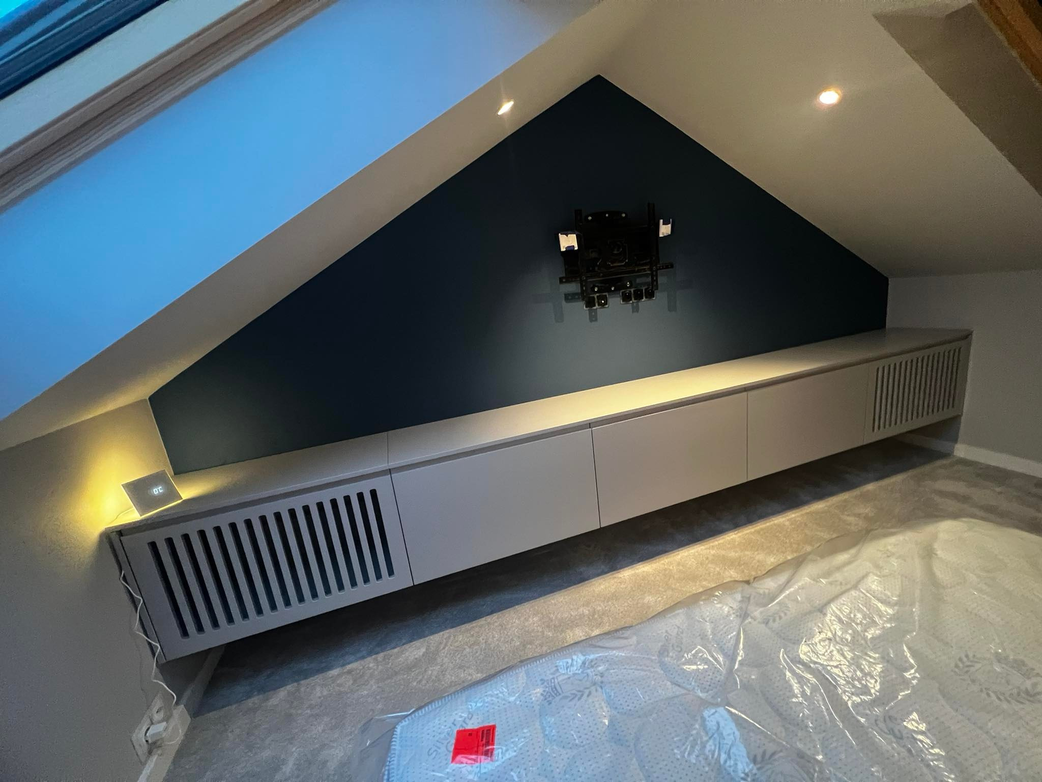 Długi, niski, szary mebel pod skosem dachu z ciemnoniebieską ścianą, na której zamontowany jest uchwyt do telewizora; na meblu mały, podświetlony zegar.