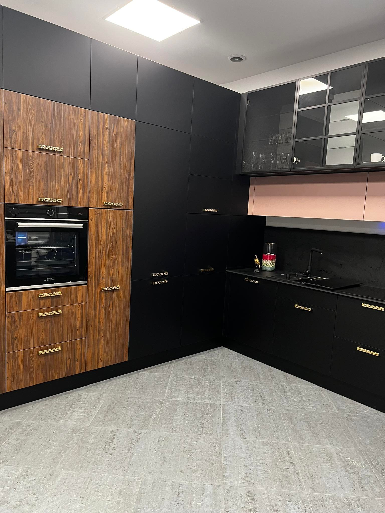 Nowoczesna kuchnia z frontami w kolorze ciemnego drewna i czerni, ozdobnymi złotymi uchwytami, szklanymi szafkami wiszącymi i szarą podłogą.