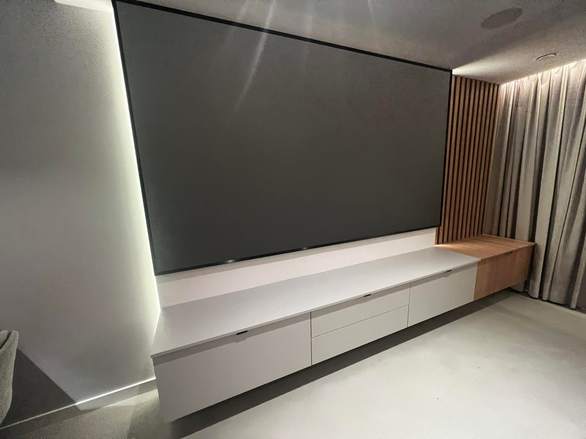 Nowoczesna szafka RTV na całej szerokości ściany w minimalistycznym salonie, z białymi frontami i drewnianymi akcentami, podwieszona pod dużym, ciemnym ekranem, oświetlona listwą LED wzdłuż ściany.