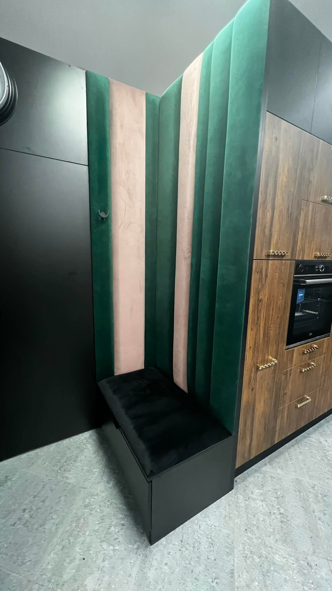 Nowoczesna kuchnia z narożnym siedziskiem obitym czarnym welurem, ściana wykończona pionowymi panelami tapicerowanymi w kolorach zieleni i pudrowego różu, sąsiadująca z szafkami kuchennymi...