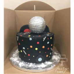 Czarny tort z motywem disco, zdobiony kolorowymi kropkami, srebrną kulą disco i czarnymi płytami winylowymi, umieszczony na srebrnej tacy w kartonowym pudełku.