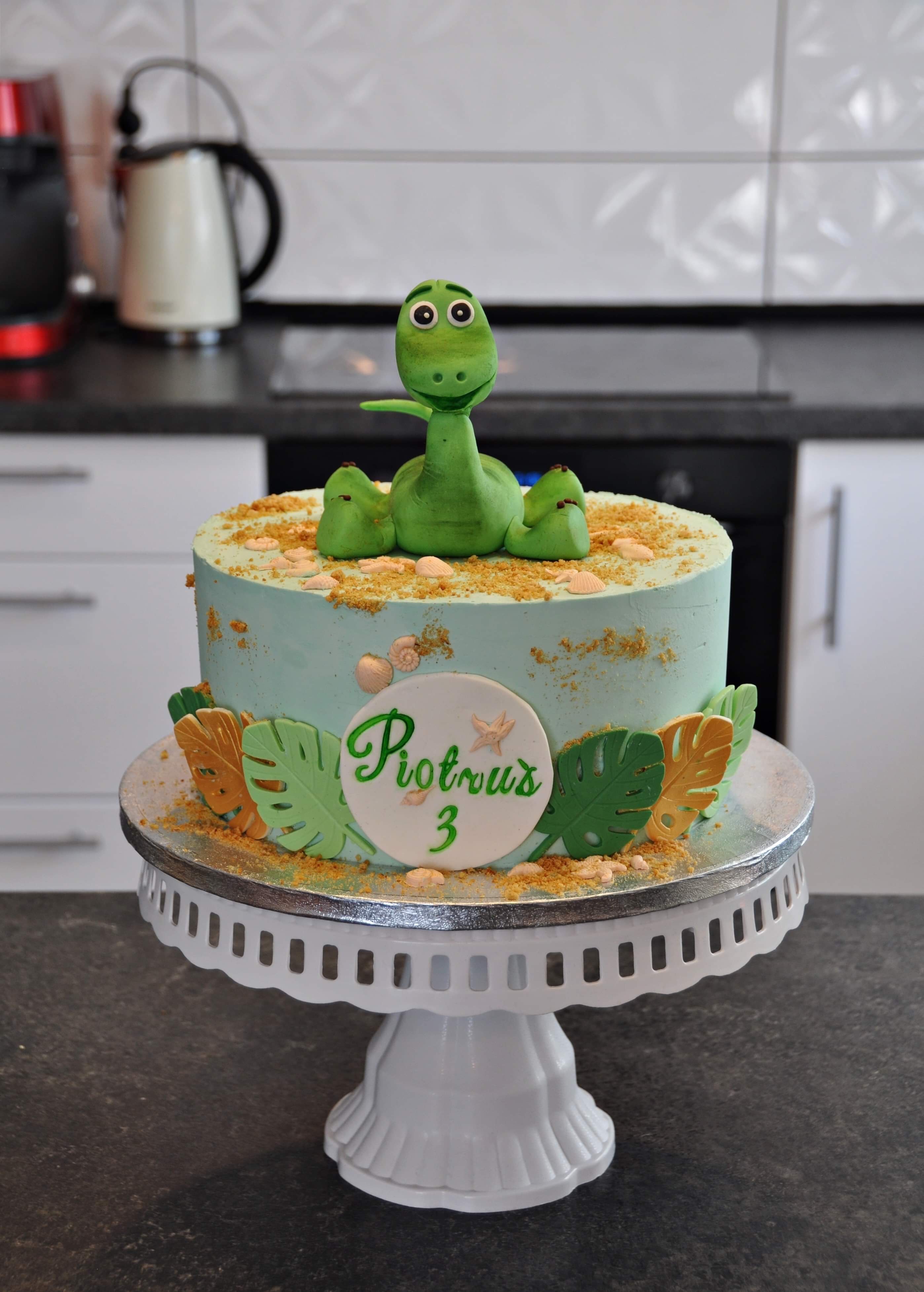Tort urodzinowy z turkusowym kremem, posypką i figurką zielonego dinozaura na białym, ozdobnym stojaku. Dekoracje z liści i napis 'Piotruś 3' na froncie.