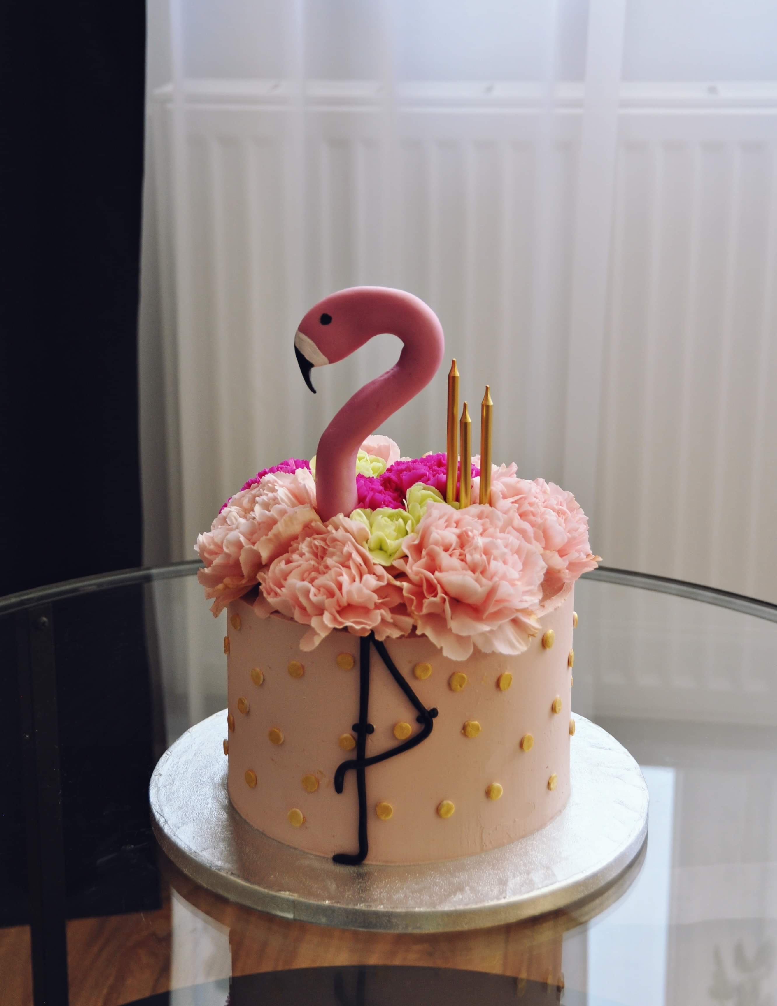 Tort z różową polewą w złote kropki, dekorowany kwiatami, figurką flaminga i złotymi świeczkami, stojący na szklanym stole.