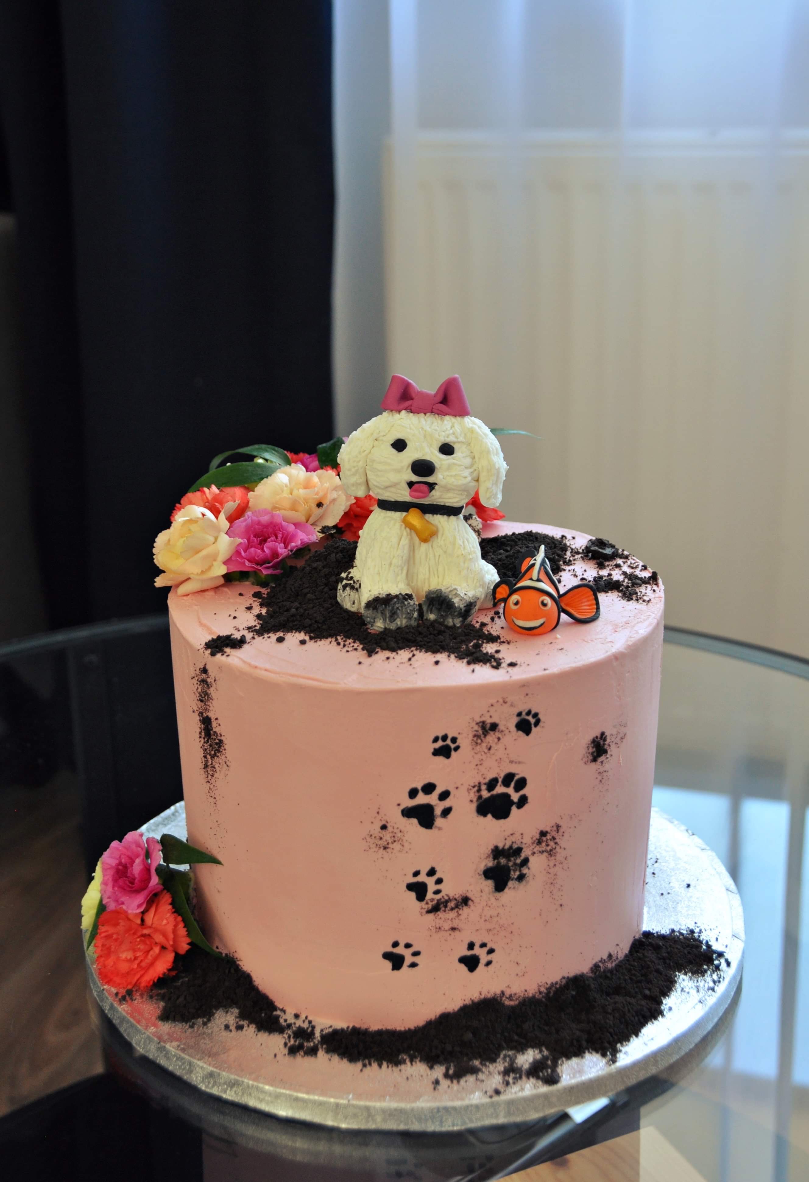 Tort w odcieniach różu z dekoracją w postaci psa z kokardą, rybki Nemo, śladów łap i kwiatów.