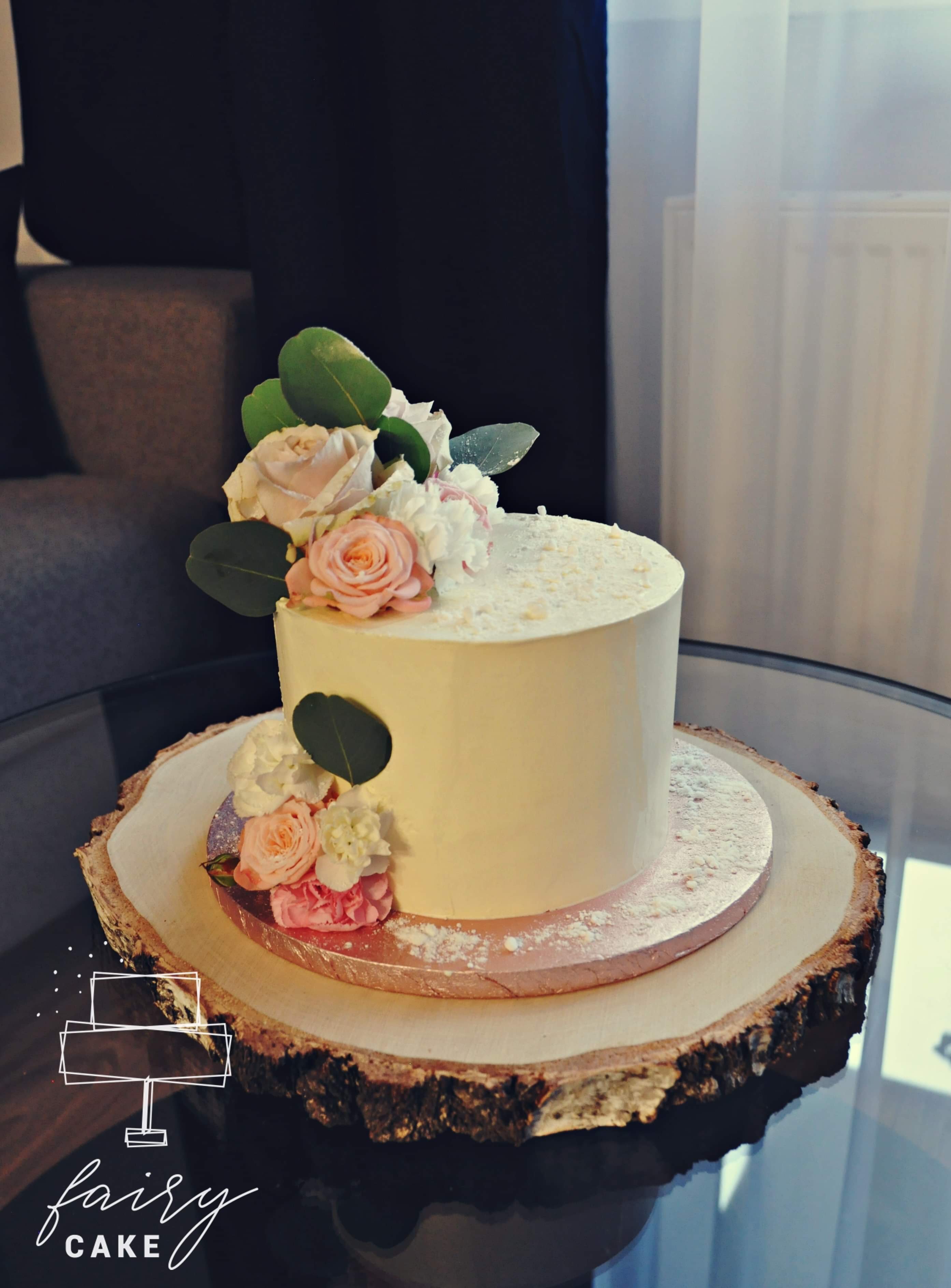 Dwupoziomowy tort z białym kremem, ozdobiony pastelowymi różami i zielonymi liśćmi, umieszczony na drewnianej podstawie imitującej plaster drzewa.