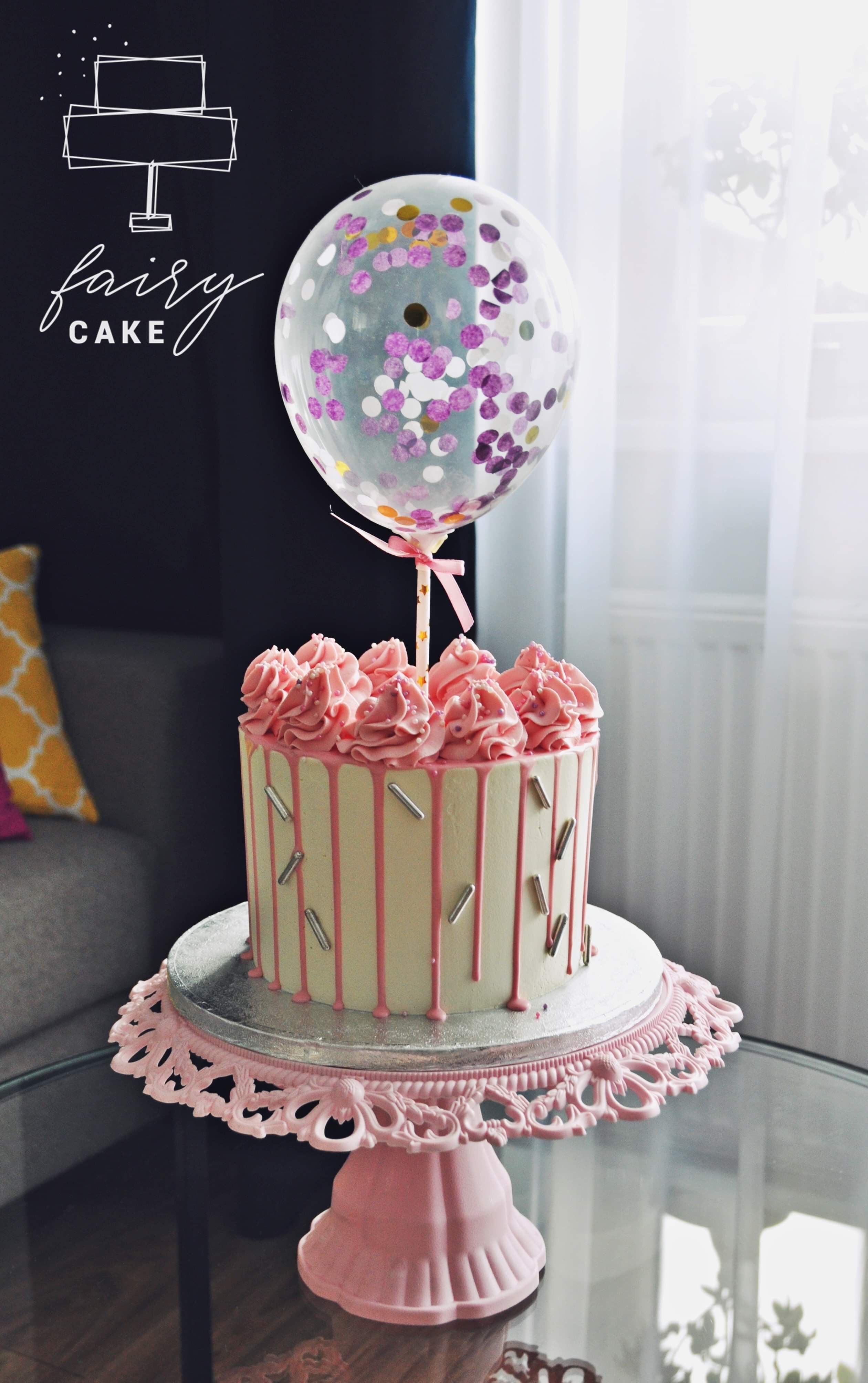 Tort urodzinowy z różową polewą, srebrnymi posypkami i balonem wypełnionym konfetti, na ozdobnym różowym stojaku.