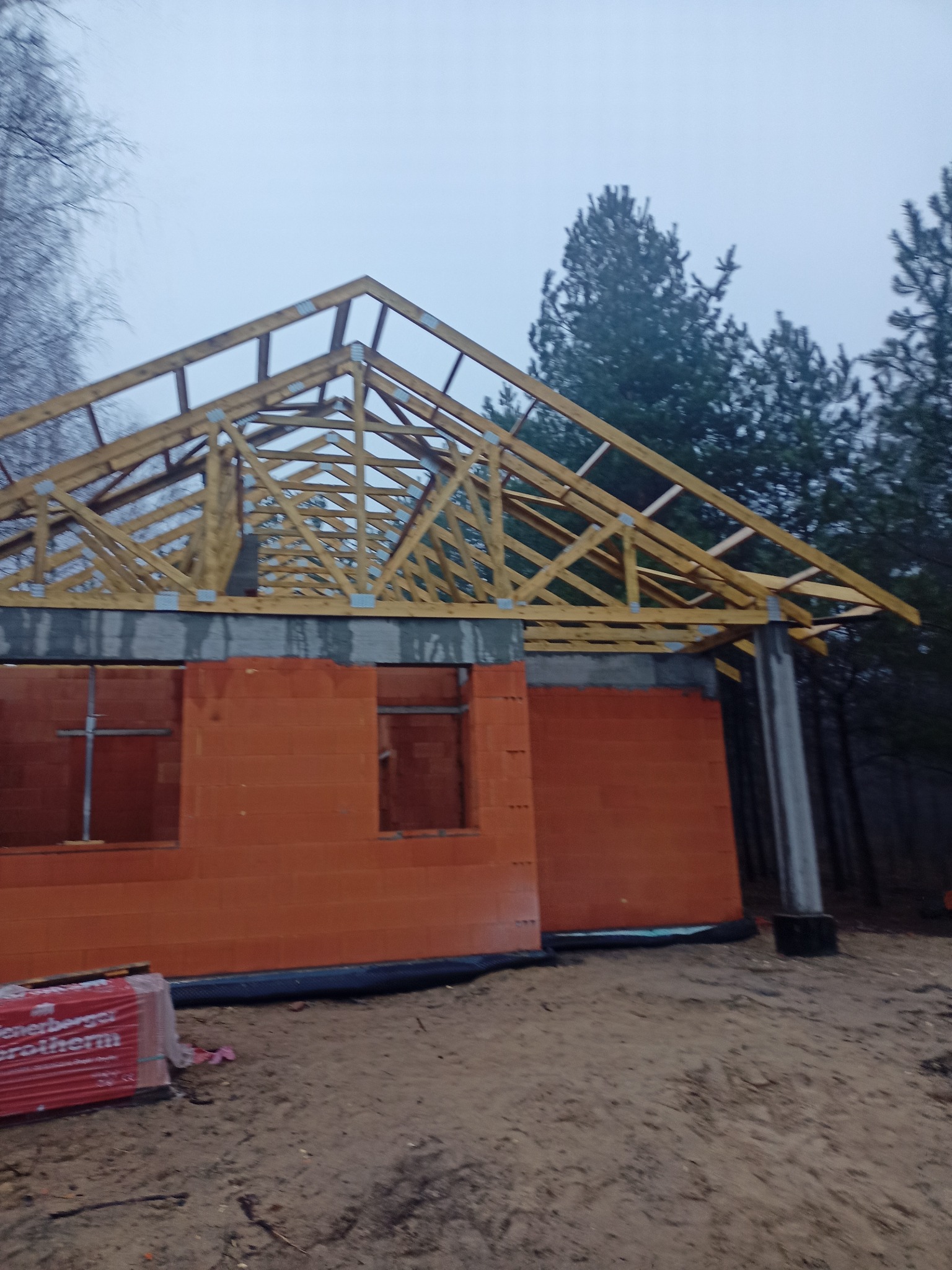 Budowa domu z czerwonej cegły Porotherm, widoczna konstrukcja dachu z drewna, betonowe słupy. Na pierwszym planie paleta cegieł. Pochmurny dzień.