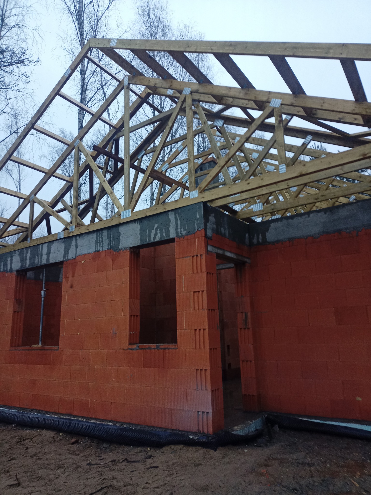 Wznoszenie domu z czerwonej cegły: ściany parteru z otworami okiennymi i drewniana konstrukcja dachu w trakcie budowy. Widok z dołu na pochmurne niebo.