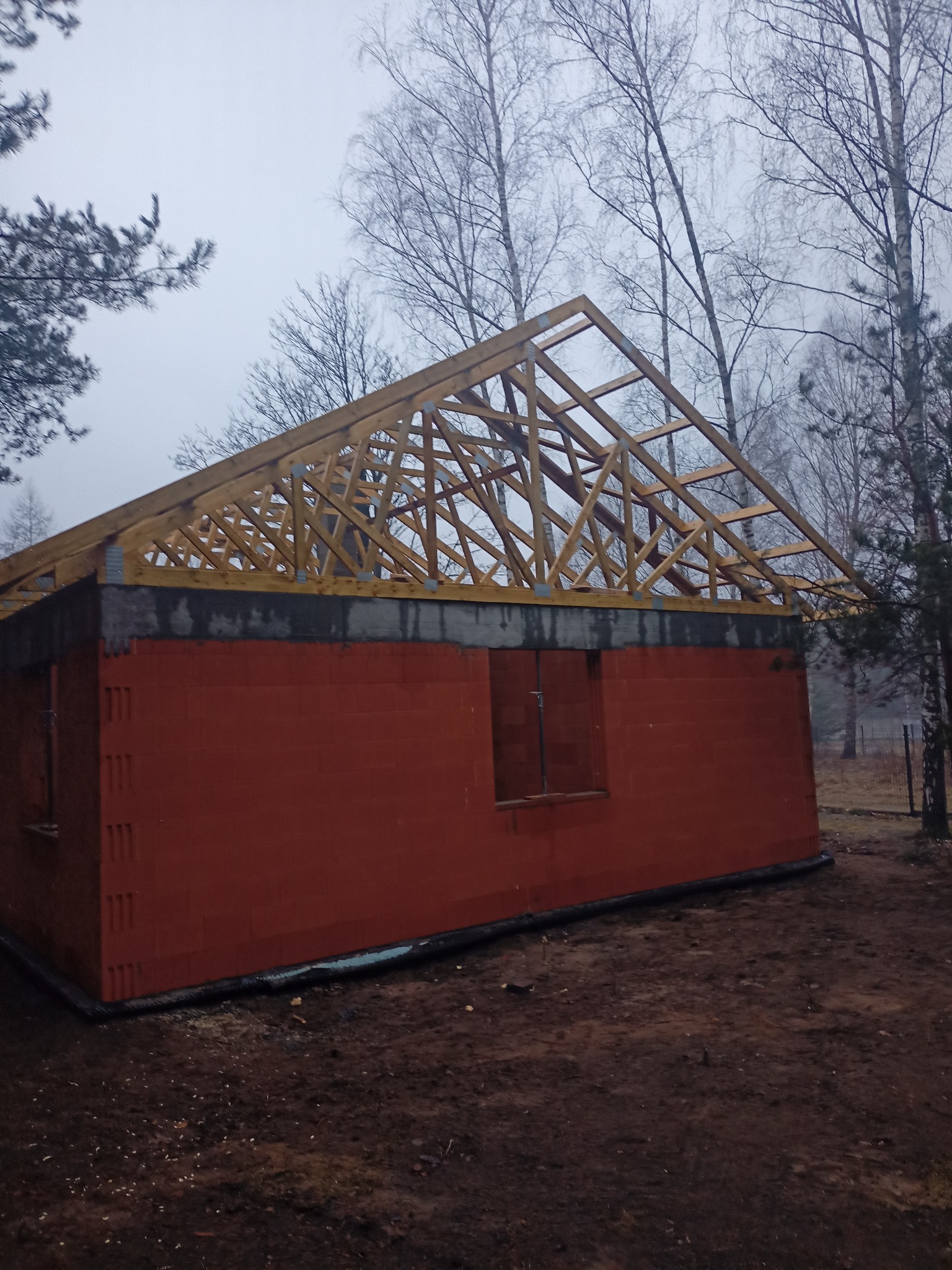 Czerwony dom w trakcie budowy z drewnianą konstrukcją dachu na tle pochmurnego nieba i drzew bez liści. Widoczny mur z pustaków i elementy więźby dachowej.
