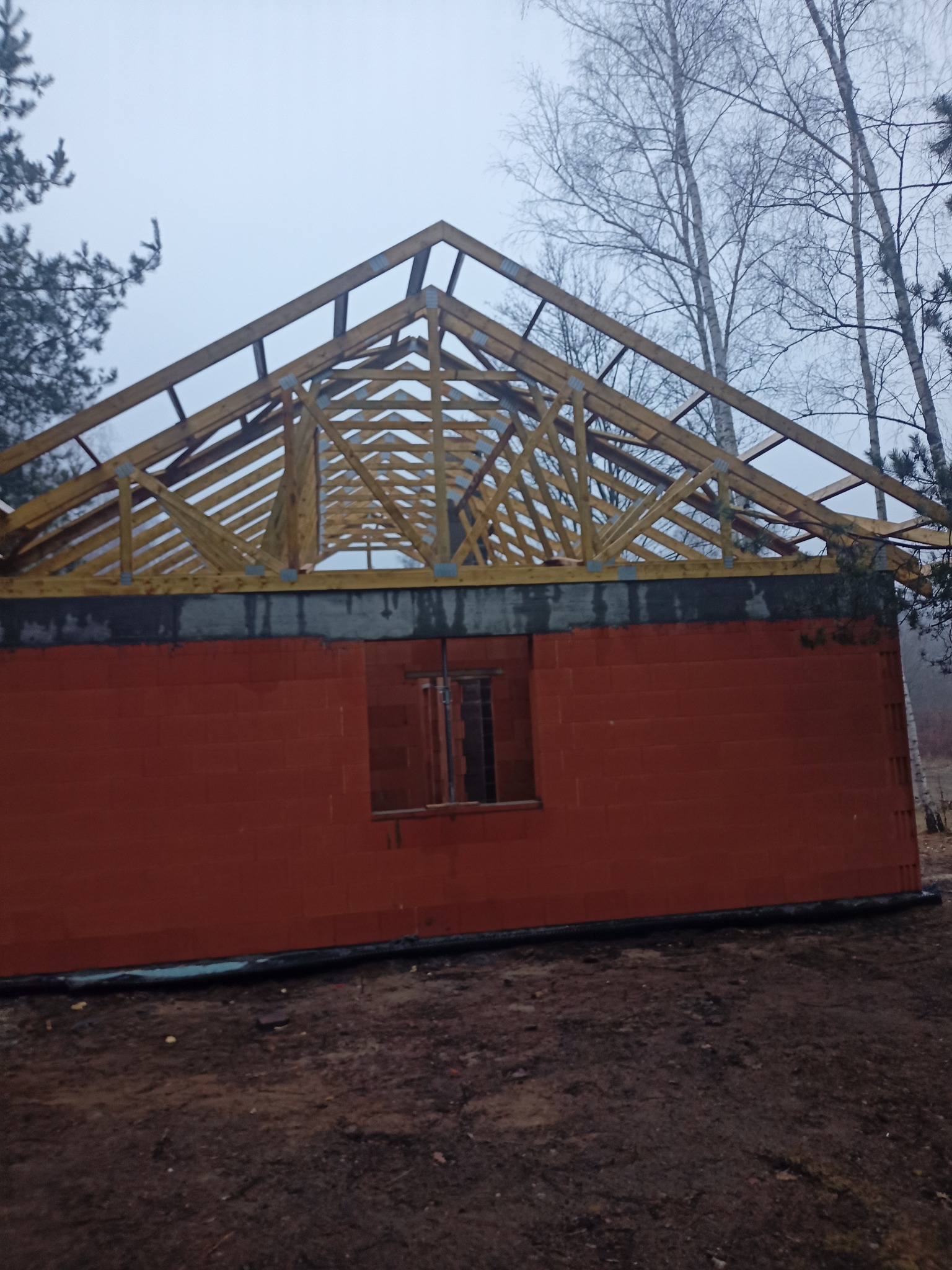Ceglany budynek w trakcie budowy z drewnianą konstrukcją dachu na tle pochmurnego nieba i drzew. Widoczny fragment okna i fundamentów.
