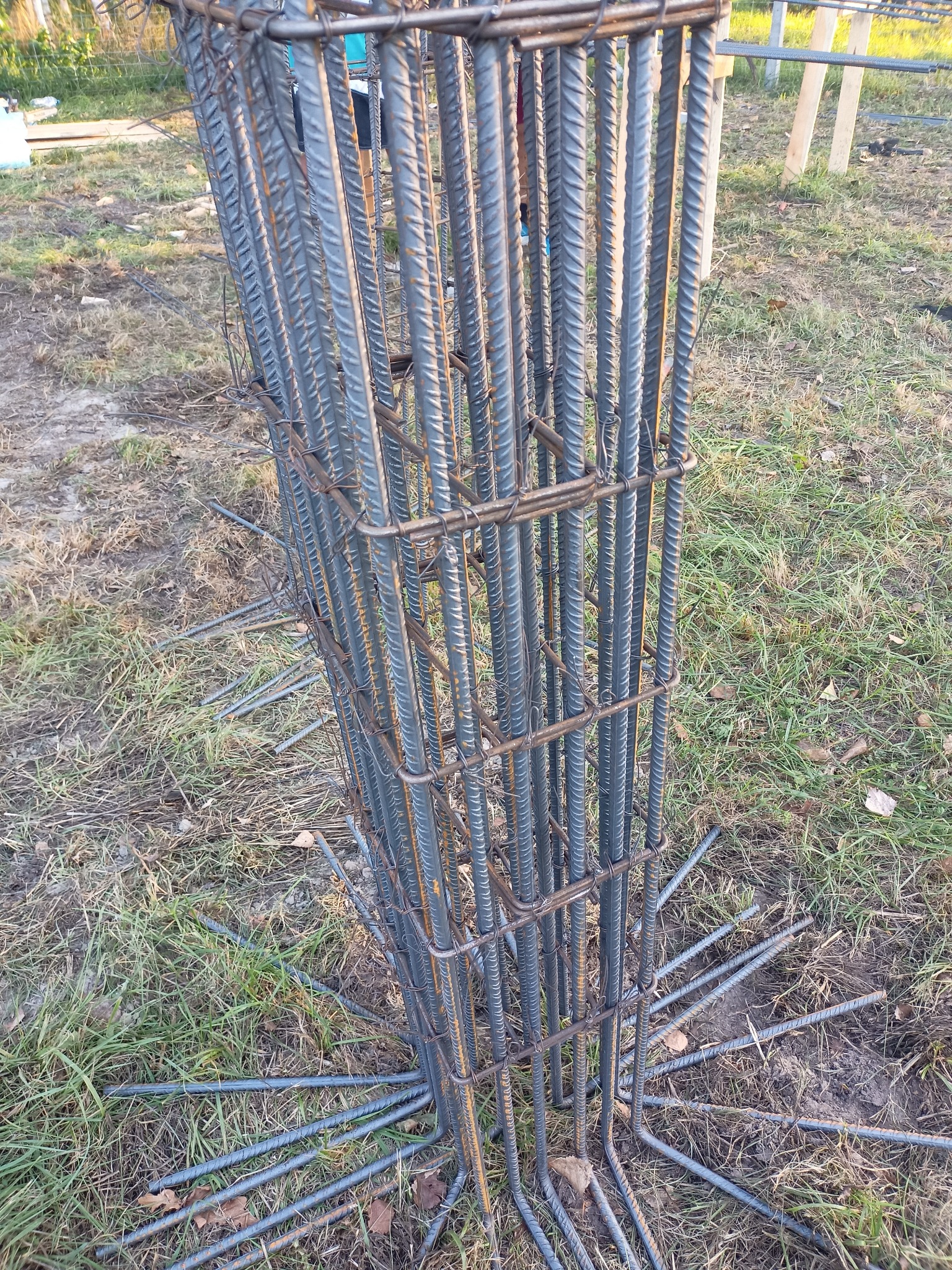 Zbrojenie kolumny fundamentowej z wystającymi prętami, przygotowane do zalania betonem na trawiastym podłożu. Widoczne wiązania drutem.