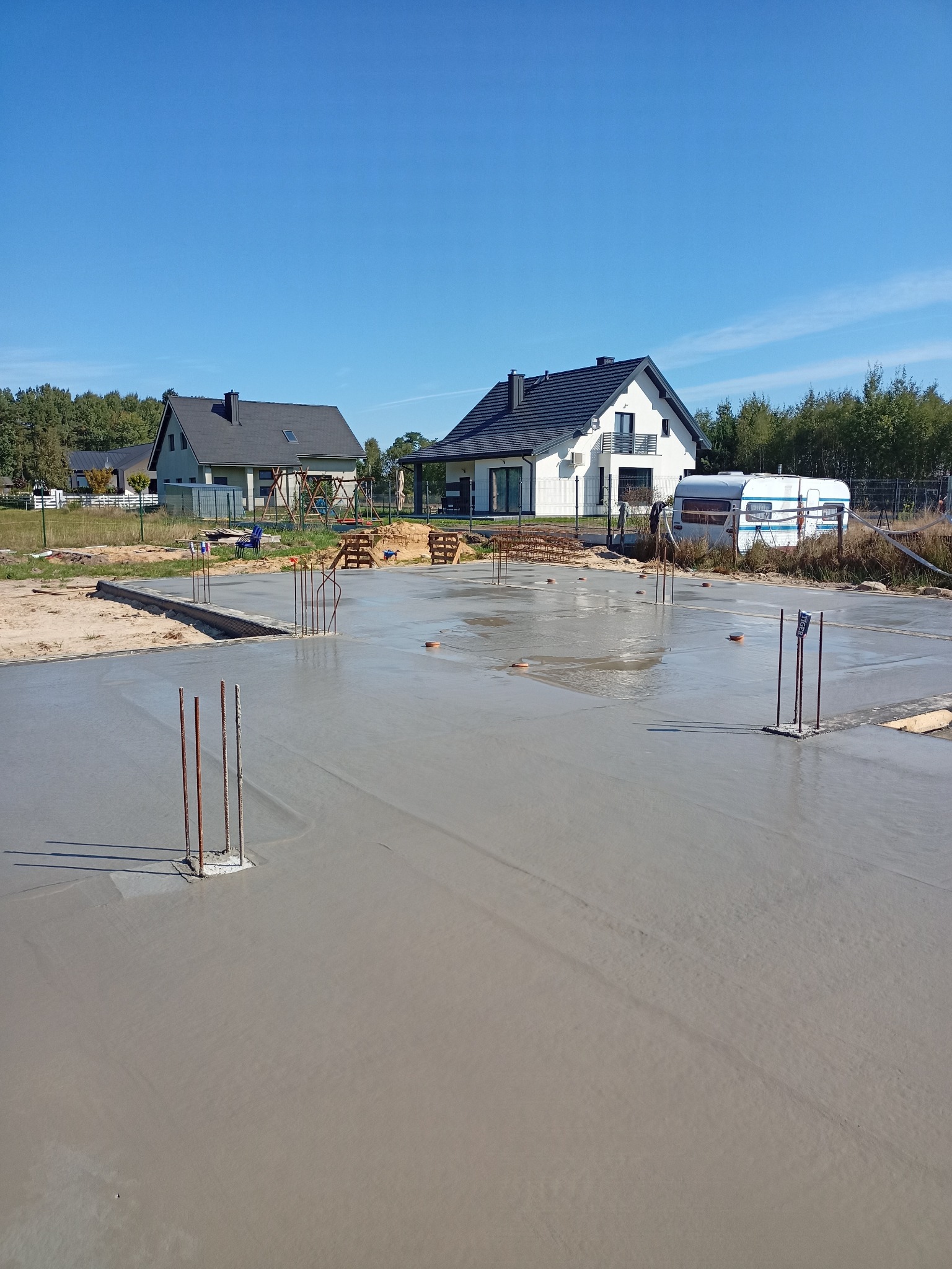 Wylany fundament pod dom jednorodzinny z wystającymi prętami zbrojeniowymi. W tle dwa domy w różnej fazie budowy i przyczepa kempingowa. Błękitne niebo.