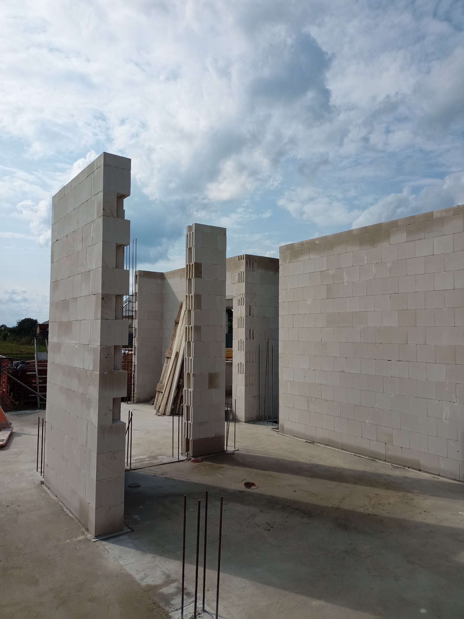 Wznoszenie ścian z betonu komórkowego na fundamencie. Widoczne zbrojenia i fragmenty deskowania. W tle błękitne niebo z chmurami.