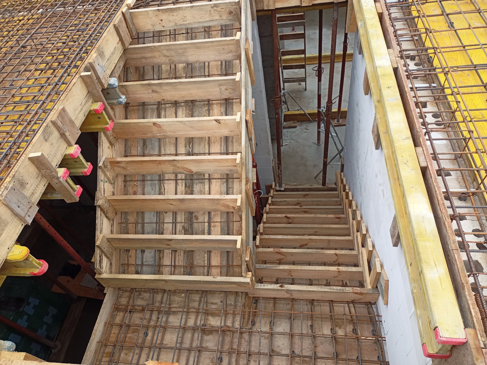 Szalunek schodów zbrojonych, widok z góry. Drewniane stopnie i metalowe pręty tworzą konstrukcję. Żółte elementy szalunku i białe bloczki w tle.