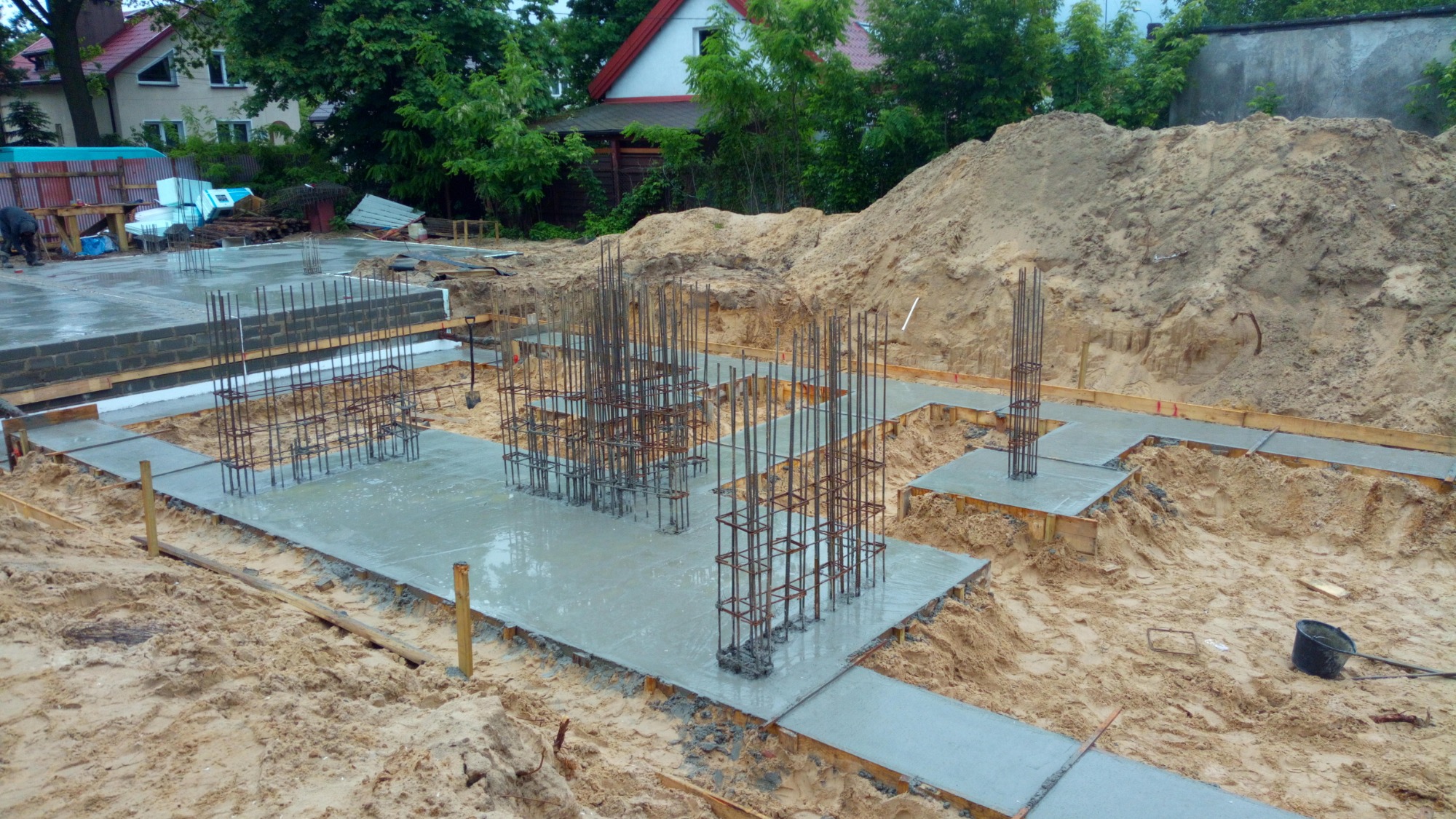 Wylane fundamenty z wystającymi prętami zbrojeniowymi, widok z góry na plac budowy, w tle dom jednorodzinny i sterta ziemi.