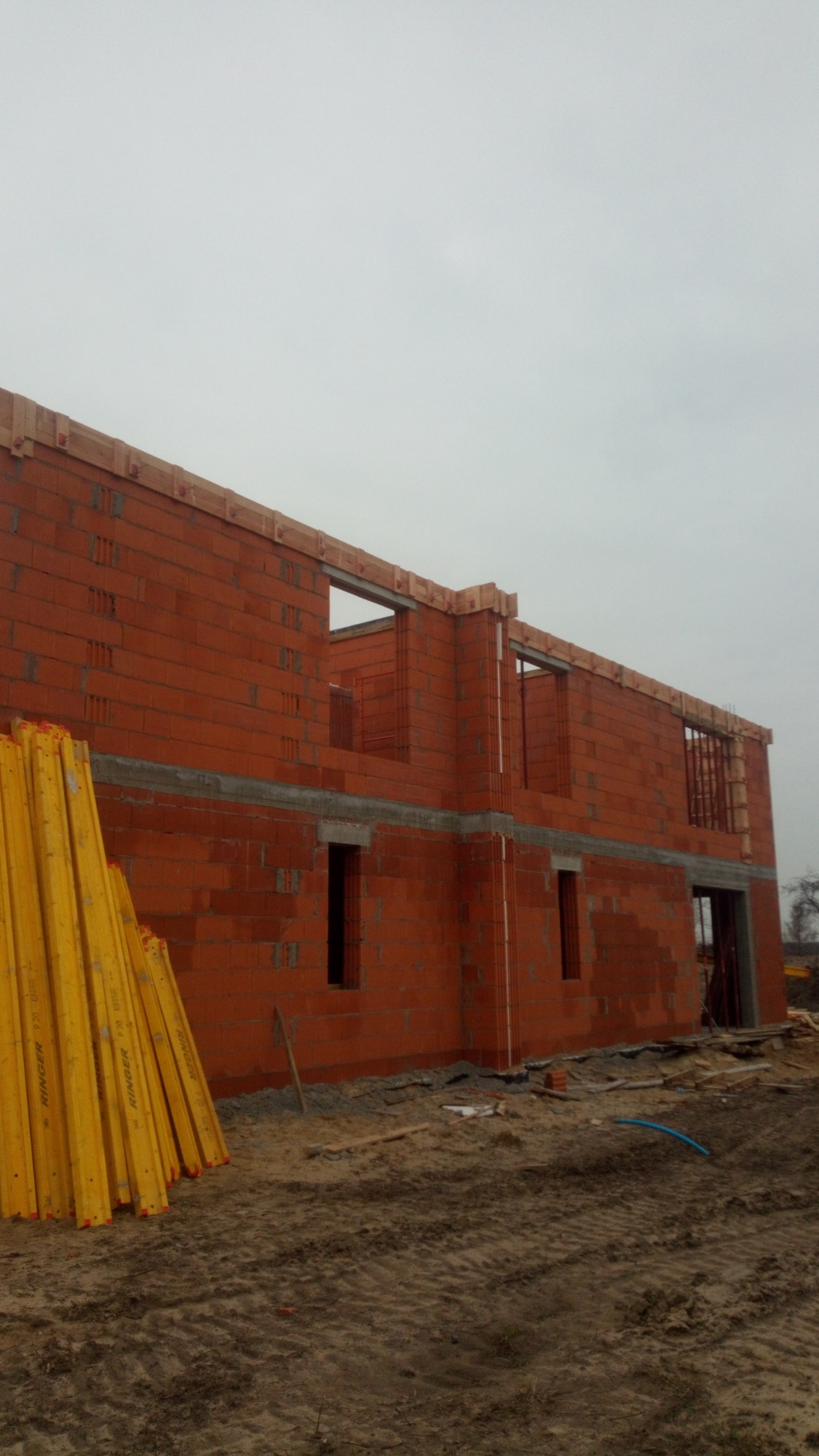 Surowy stan dwukondygnacyjnego budynku z czerwonej cegły, widoczne otwory okienne i drzwiowe, żółte stemple budowlane leżące obok, ziemisty teren budowy.