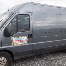 Szary samochód dostawczy z naklejką reklamową wypożyczalni skuterów elektrycznych na boku, zaparkowany na żwirowym podłożu, z widocznym budynkiem i stertą opon w tle.