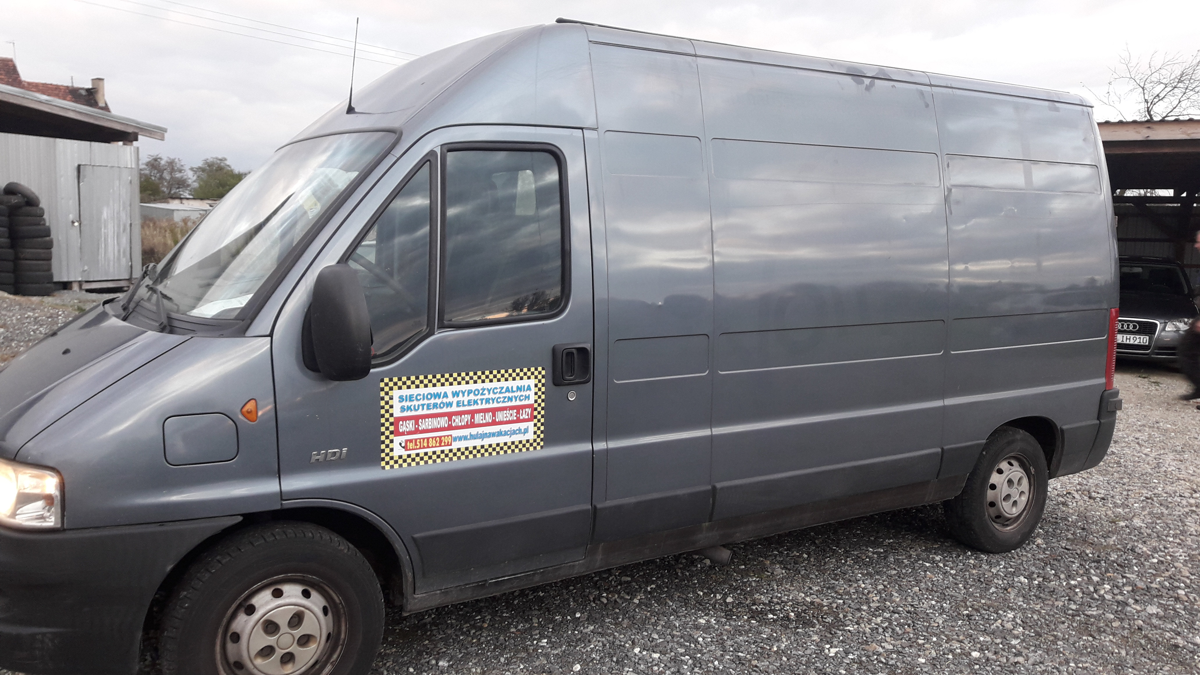 Szary samochód dostawczy z naklejką reklamową wypożyczalni skuterów elektrycznych na boku, zaparkowany na żwirowym podłożu, z widocznym budynkiem i stertą opon w tle.