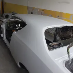 Nadwozie klasycznego samochodu w trakcie lakierowania, pokryte szarym podkładem, widoczny fragment wnętrza i koło z oponą Goodyear, wnętrze warsztatu w tle.