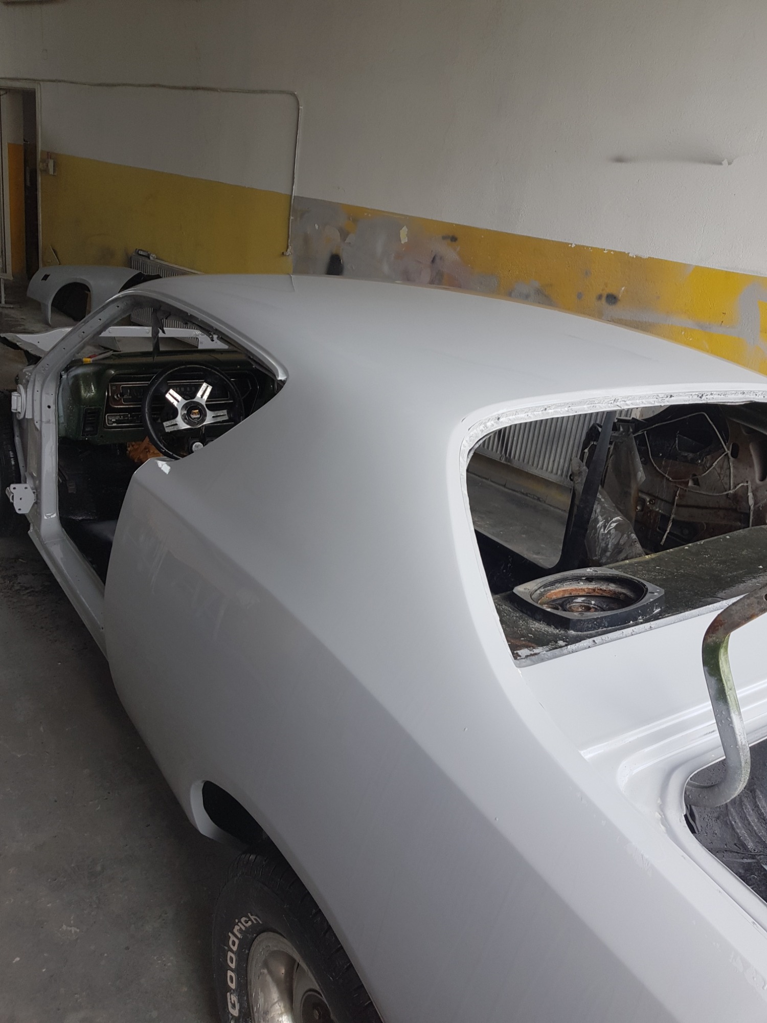 Nadwozie klasycznego samochodu w trakcie lakierowania, pokryte szarym podkładem, widoczny fragment wnętrza i koło z oponą Goodyear, wnętrze warsztatu w tle.