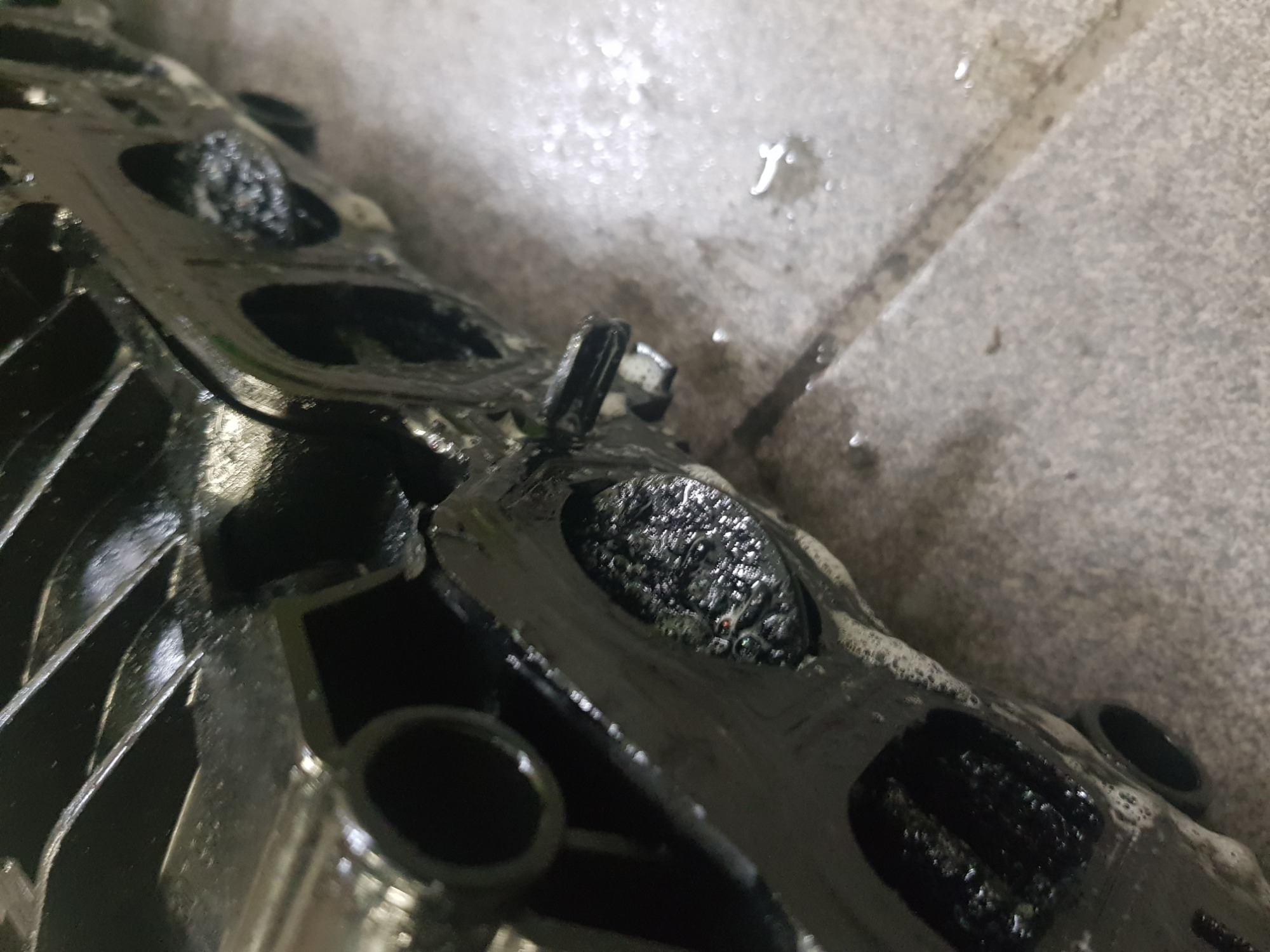 Zdemontowany, bardzo brudny kolektor dolotowy silnika samochodowego, pokryty olejem i osadami, leżący na betonowej podłodze warsztatu.