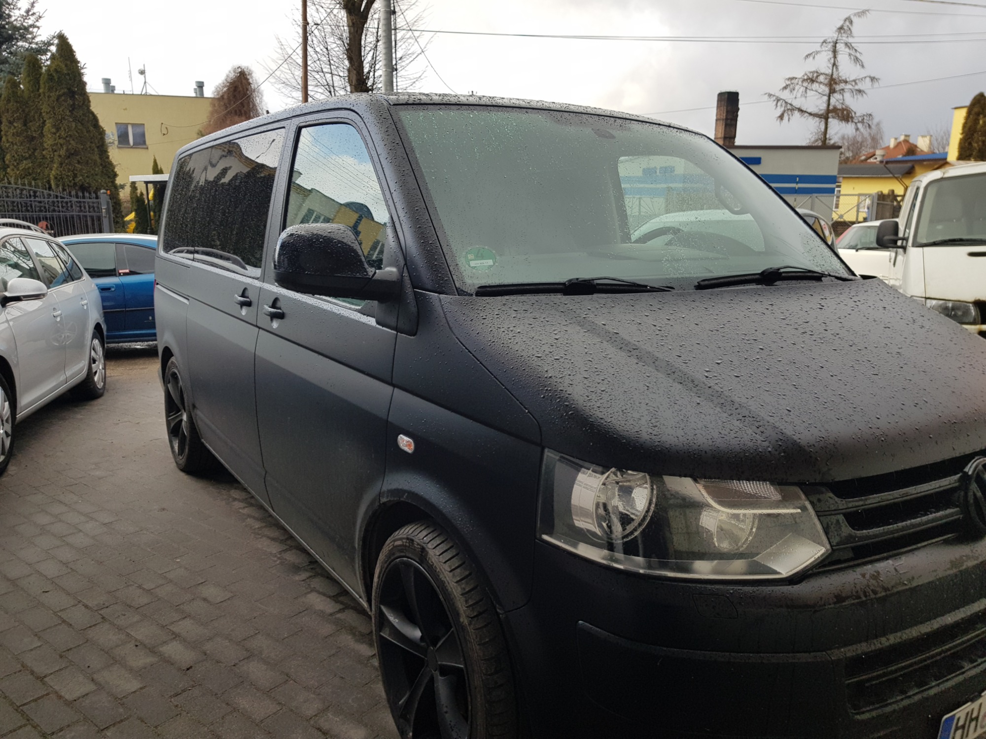 Czarny, matowy Volkswagen Transporter T5 z przyciemnionymi szybami, pokryty kroplami deszczu, zaparkowany na brukowanej powierzchni.
