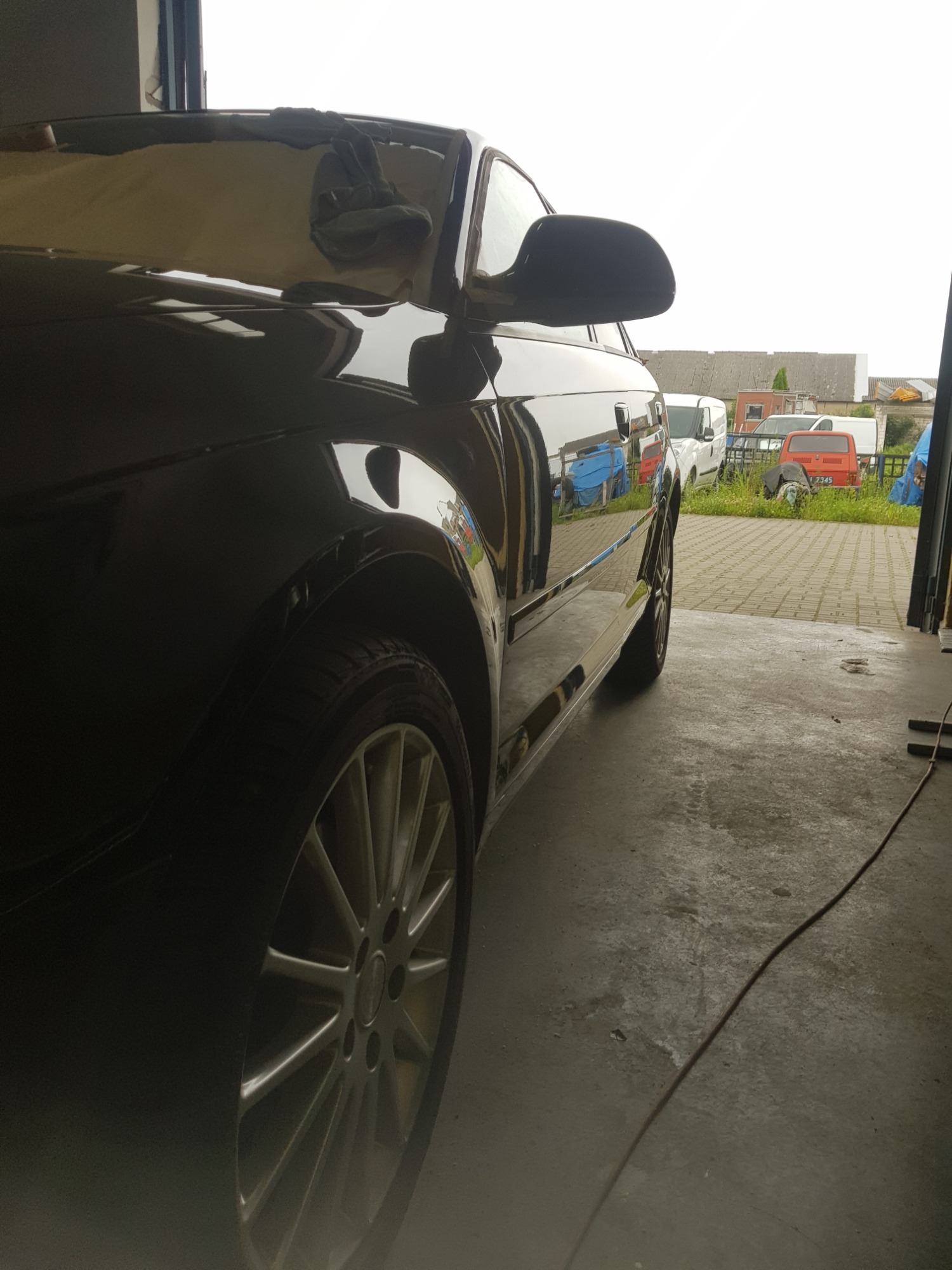 Błyszczący, czarny samochód po polerowaniu, widok z boku, z widocznym odbiciem otoczenia na karoserii i felgami aluminiowymi. W tle inne pojazdy i budynki.