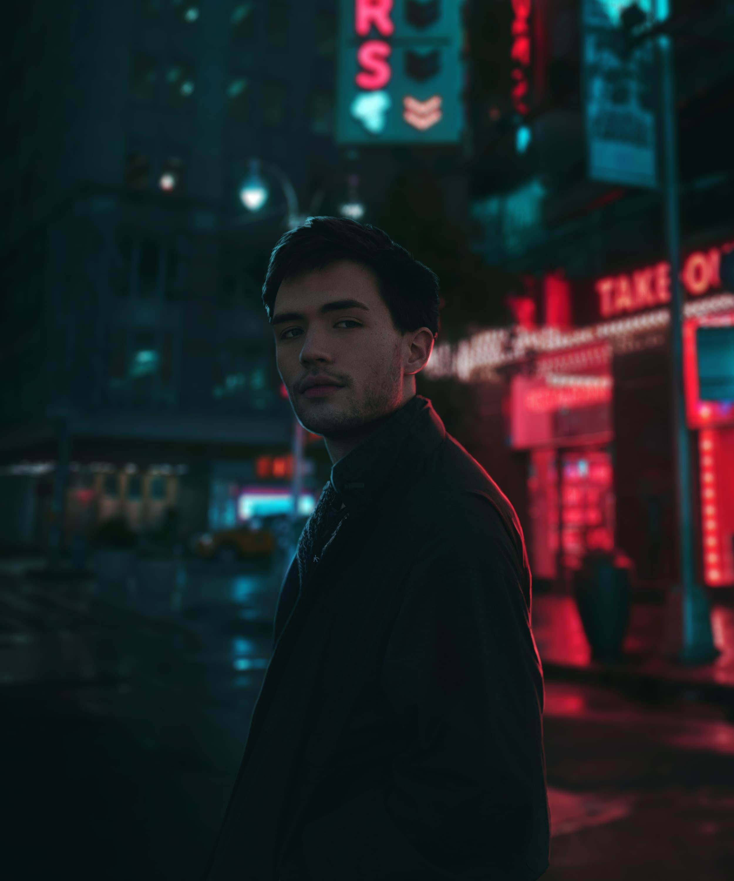 Młody mężczyzna w ciemnej kurtce na tle neonowych świateł miejskich, z rozmazanym szyldem 'TAKE OUT' w czerwieni i niebieskim szyldem w tle, sugerujący nocną scenerię i potencjalną sesję zdjęciową.