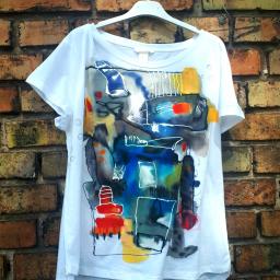 mala galeryjka - Biała koszulka damska z abstrakcyjnym, ręcznie malowanym wzorem w odcieniach niebieskiego, czerwonego, żółtego i szarego, zawieszona na wieszaku na tle ceglanej ściany.