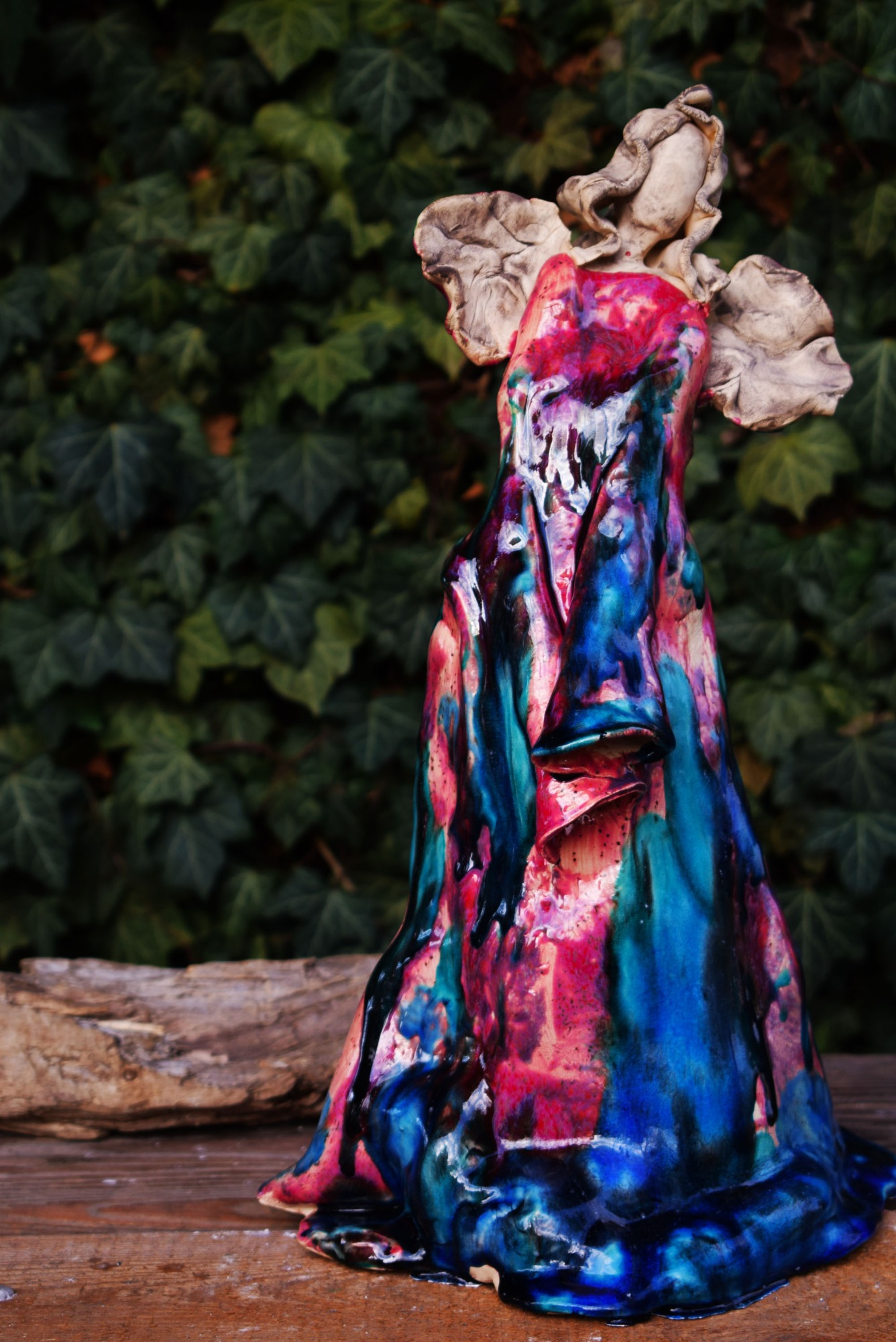 Ceramiczna figurka anioła w jaskrawych kolorach (róż, turkus, niebieski) z efektem spływającej glazury, na tle zielonego bluszczu i kawałka drewna.