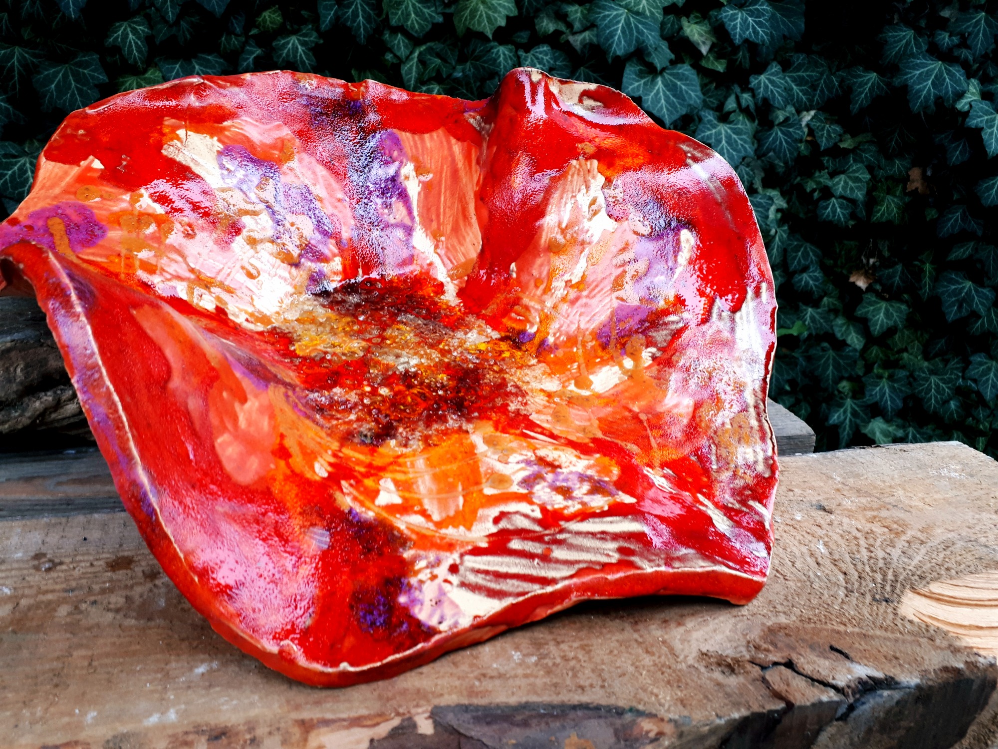 Ręcznie formowana, szkliwiona misa ceramiczna w odcieniach czerwieni, pomarańczy i bieli, umieszczona na surowym drewnianym blacie z zielonym bluszczem w tle.