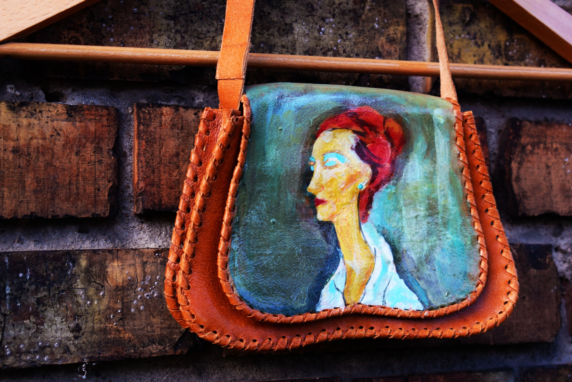 Ręcznie malowana torebka damska z brązowej skóry, wisząca na drewnianym wieszaku na tle ceglanej ściany, przedstawiająca portret kobiety w stylu vintage z turkusowymi oczami.