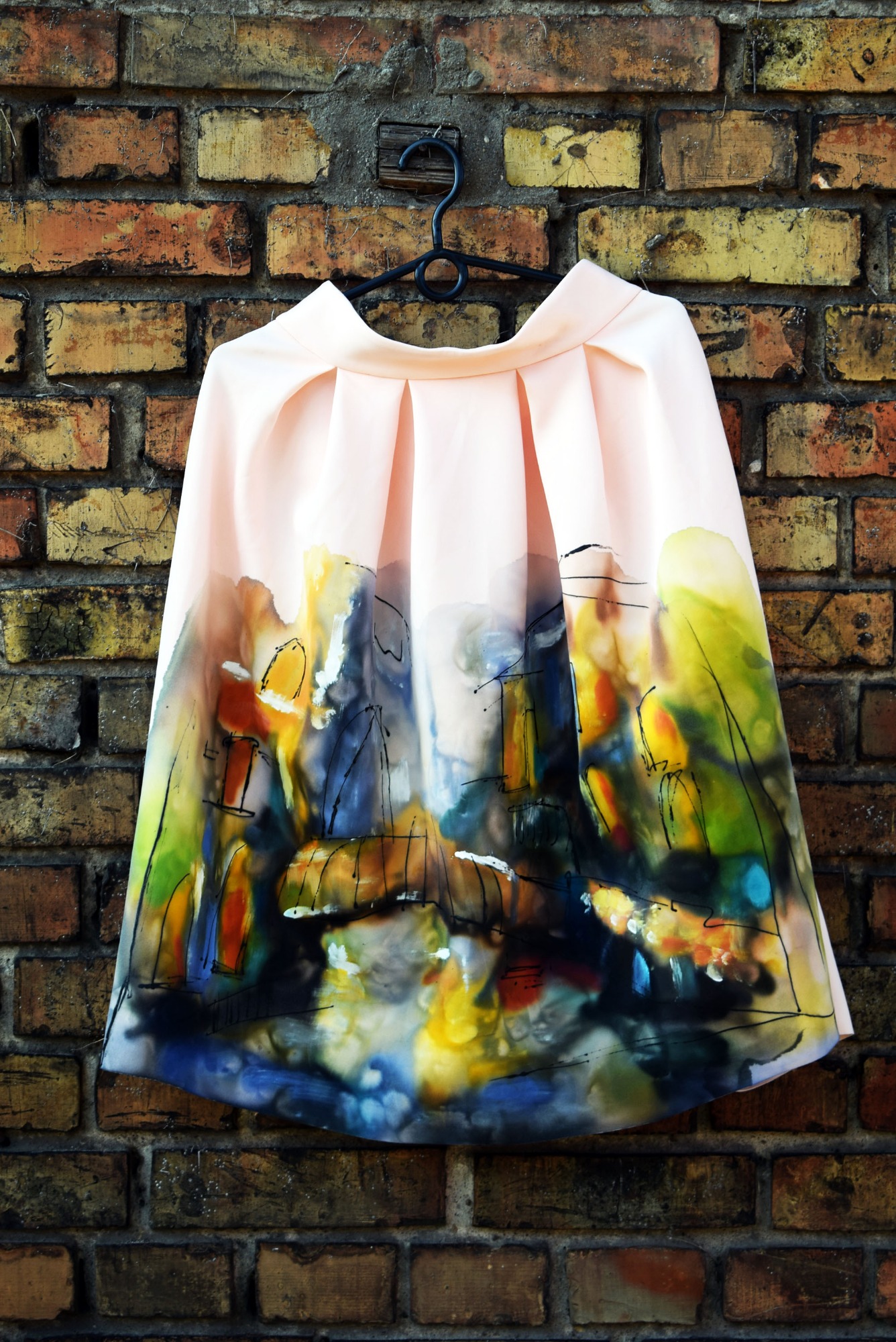 Jasnoróżowa spódnica z plisami i abstrakcyjnym nadrukiem krajobrazu, wisząca na wieszaku na tle ceglanej ściany.