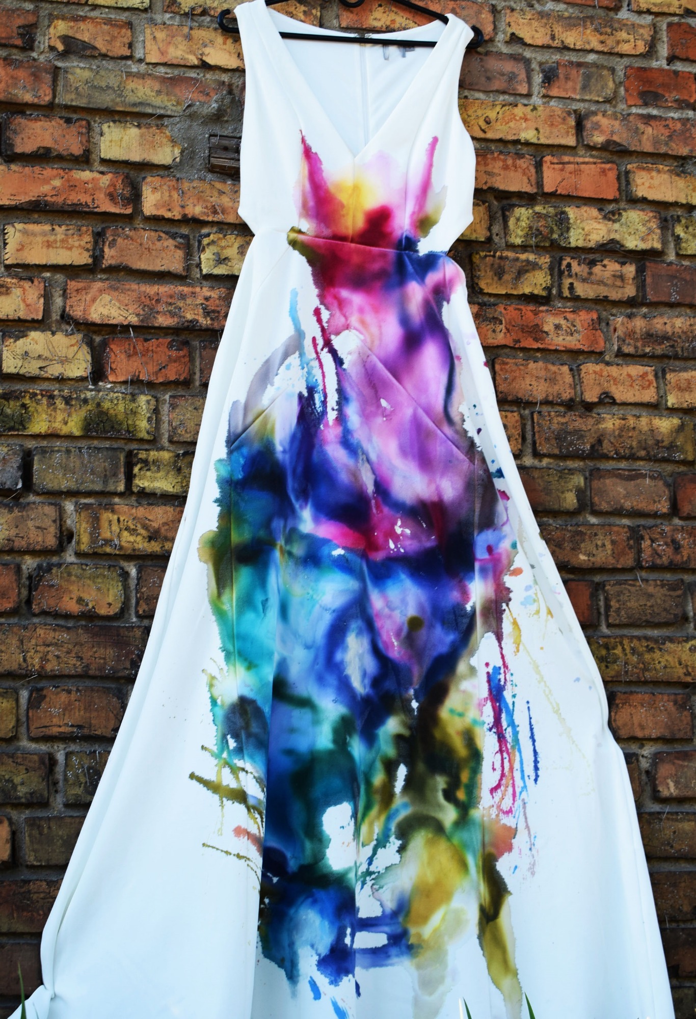 Biała suknia wieczorowa z abstrakcyjnymi, kolorowymi nadrukami w odcieniach niebieskiego, fioletu, różu i żółci, zawieszona na wieszaku na tle ściany z czerwonej cegły.