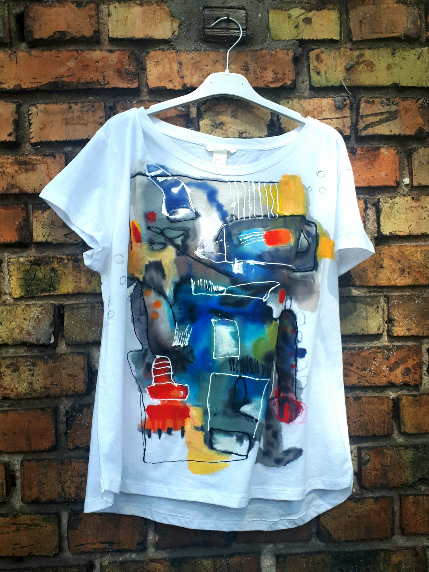Biała koszulka damska z abstrakcyjnym, ręcznie malowanym wzorem w odcieniach niebieskiego, czerwonego, żółtego i szarego, zawieszona na wieszaku na tle ceglanej ściany.