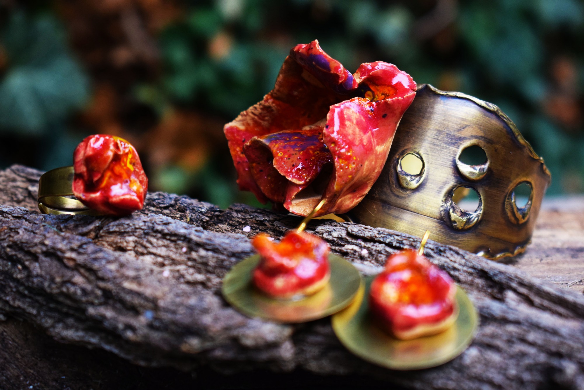 Ręcznie robiona biżuteria artystyczna: pierścionek z czerwonym oczkiem, kolczyki na złotych blaszkach i ażurowa bransoletka z mosiądzu, ułożone na kawałku kory drzewa na tle zieleni.