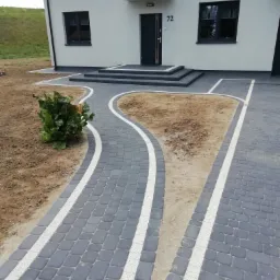 Ułożona ścieżka z szarej kostki brukowej z białymi pasami, prowadząca do domu z numerem 72 i schodami wejściowymi, obok rabata z roślinnością.