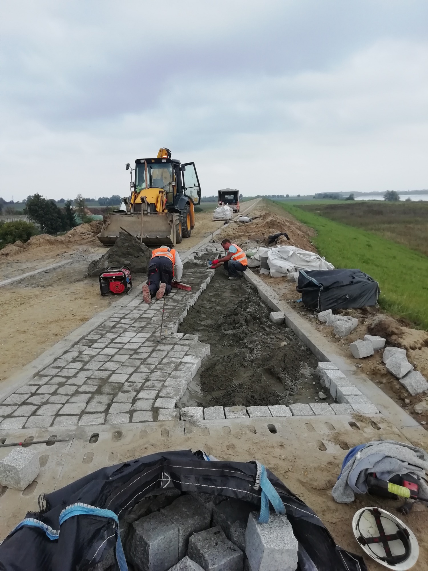 Układanie chodnika z kostki granitowej na poboczu drogi, widoczny żółty traktor i dwóch pracowników w kamizelkach odblaskowych, jeden klęczy i zamiata piasek z kostki, drugi układa kolejne kostki.