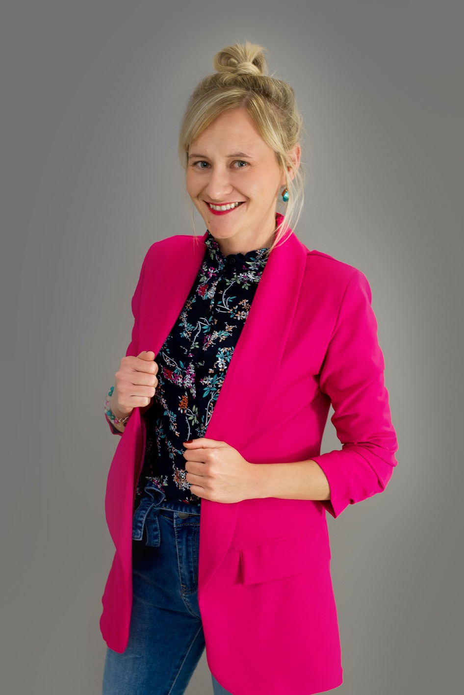 Portret uśmiechniętej kobiety w różowym żakiecie i kwiecistej bluzce na szarym tle.
