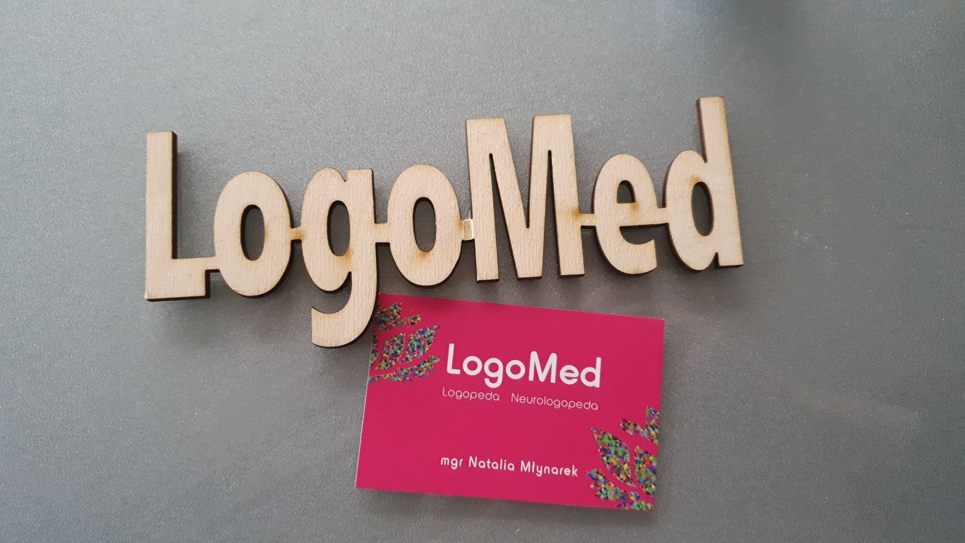 Drewniany napis 'LogoMed' nad różową wizytówką z tym samym napisem i danymi kontaktowymi mgr Natalii Młynarek, na szarym tle.