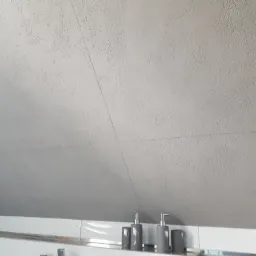 Szary, teksturowany sufit pod skosem w łazience, z widocznymi łączeniami płyt kartonowo-gipsowych i akcesoriami łazienkowymi na półce poniżej.